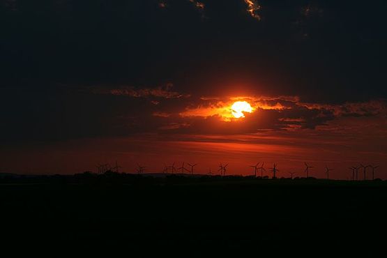 Foto zeigt Sonnenuntergang, der glutrote Feuerball der Sonne bricht durch die Wolken und strahlt sein rotes Licht auf die WindkraFoto zeigt Sonnenuntergang, der glutrote Feuerball der Sonne bricht durch die Wolken und strahlt sein rotes Licht auf die Wind