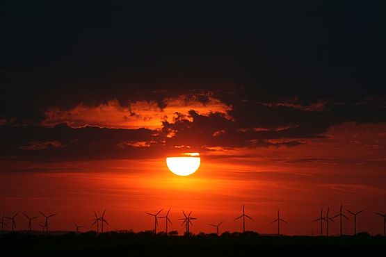 Foto zeigt Sonnenuntergang, der glutrote Feuerball der Sonne sinkt unter die Wolken und strahlt sein rotes Licht auf die Windkraftanlagen der Parndorfer Platte am Horizont