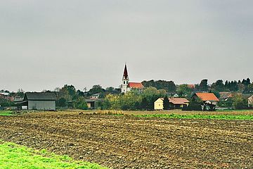 Foto zeigt Blick auf die katholische Kirche vom Bereich Wiesenäcker mit Blickrichtung Südwest, im Vordergrund ein frisch gepflügtes Feld, im Hintergrund Häuser und die Kirche